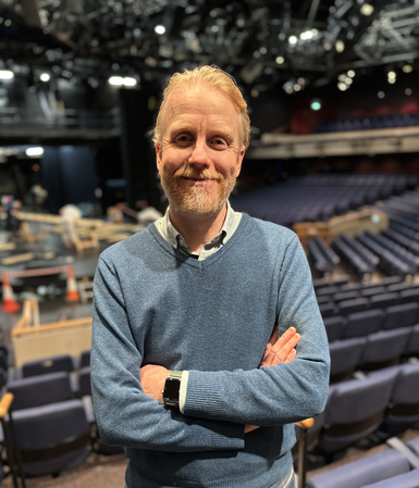 Sam Garner-Gibbons, Technical Director wears a blue jumper. He is smiling at the camera, with the CFT auditorium behind him. Lots of blue seats can be seen behind him, with the stage at the centre and lights hanging from the ceiling above.
