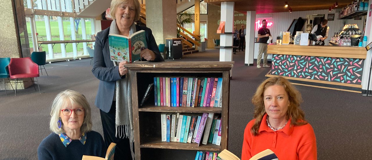 Three women sit or stand around a bookcase in the foyer of Chichester Festival Theatre. Each holds an open book and smile at the camera.