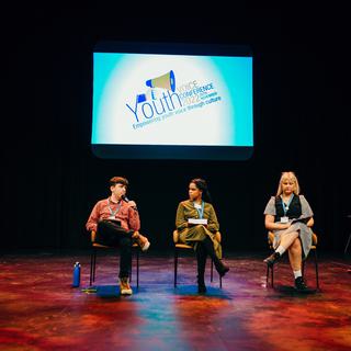 Three young people are seated on chairs with a dark backdrop behind them and a illuminated screen with the Youth Conference logo on it.