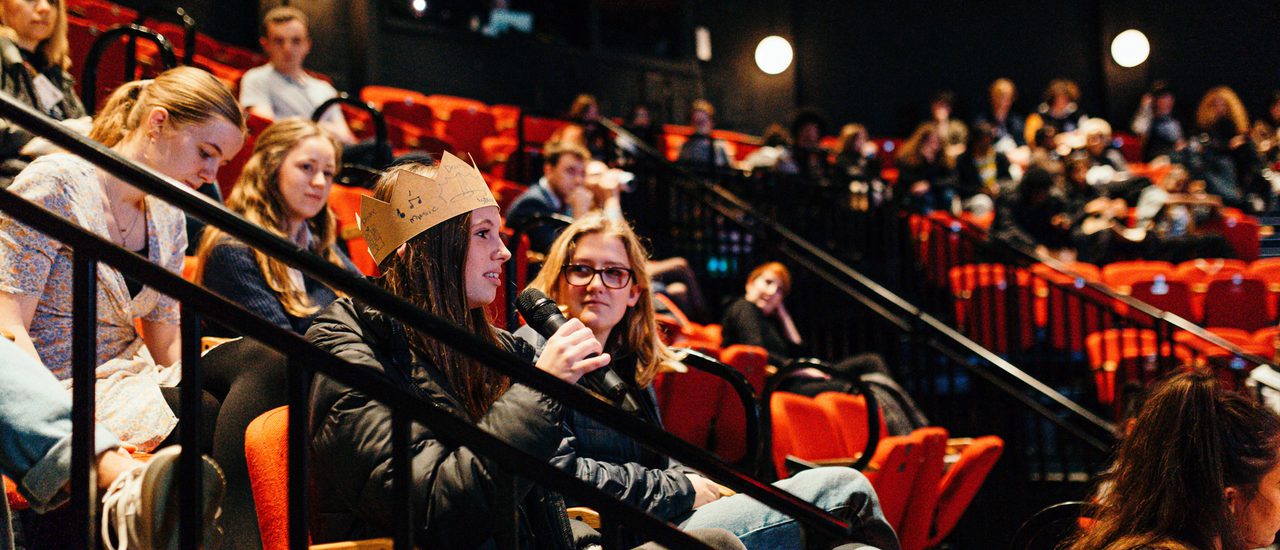 In the Minerva theatre an audience of young people are seated. A girl in the foreground is speaking into a microphone.