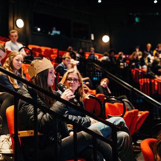In the Minerva theatre an audience of young people are seated. A girl in the foreground is speaking into a microphone.