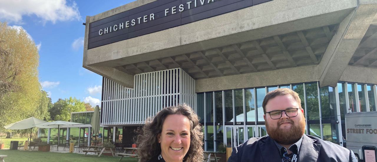 Sally and Joe stand smiling at the camera with Chichester Festival Theatre behind them. Sally is on the left with dark brown hair and waring a black and white shirt; Joe, on the right, has dark hair and a beard and is wearing black.