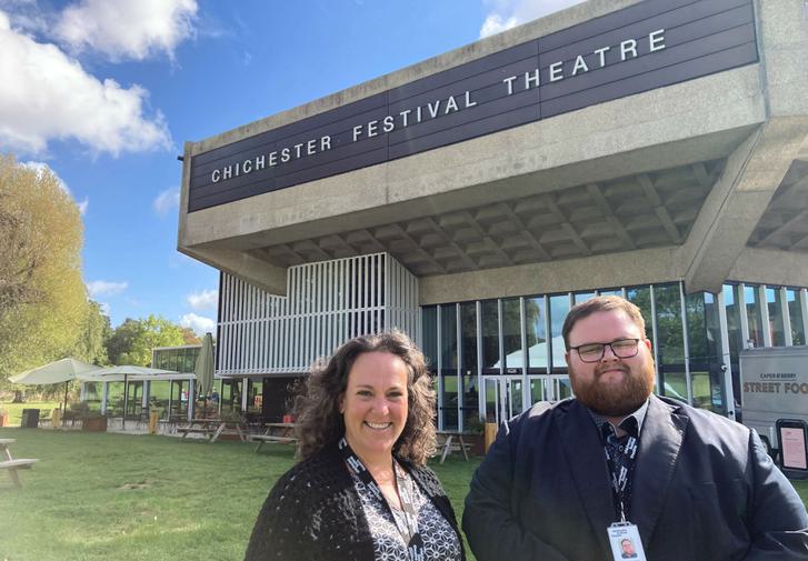 Sally and Joe stand smiling at the camera with Chichester Festival Theatre behind them. Sally is on the left with dark brown hair and waring a black and white shirt; Joe, on the right, has dark hair and a beard and is wearing black.