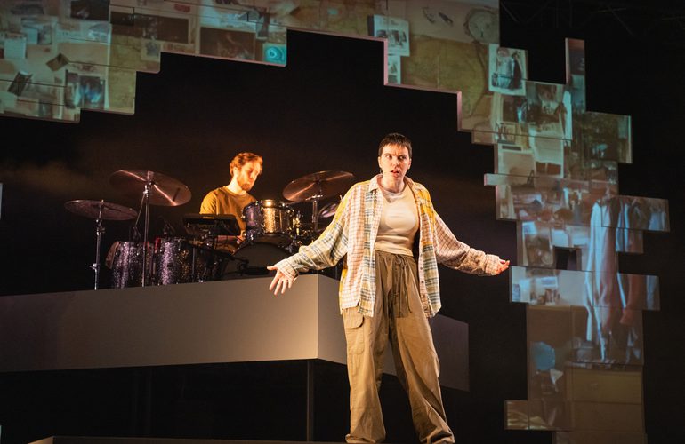 A young performer stands speaking with their arms outstretched to the sides. Behind them a drummer sits and plays a full drum kit. There is a pixelated backdrop surrounding them with distorted projections on.