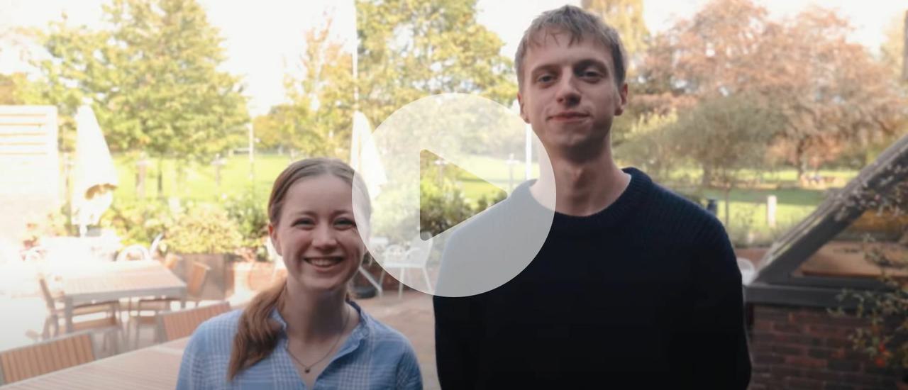 A sunny day, two people stood facing the camera smiling, Frankie is wearing a checked blue shirt and has her hair in a pony tail, George is wearing a black jumper.