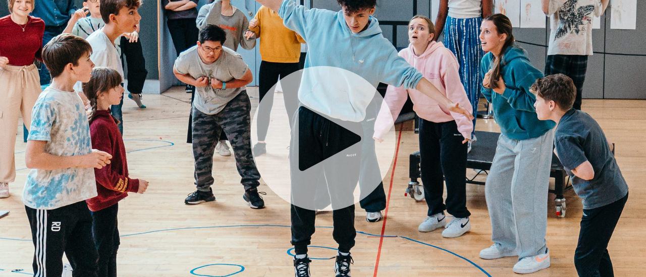 A rehearsal room full of young people. A group in the centre surround a boy who has his arms stretched out, as if he is walking on a tightrope. Others in the background are in animal poses. There is a white play button in the middle of the image,