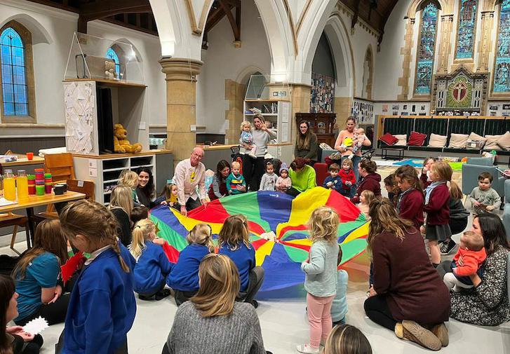 The inside of a church, a group of children and some adults gather around a large piece of coloured fabric, all engaged in what an adult is telling them