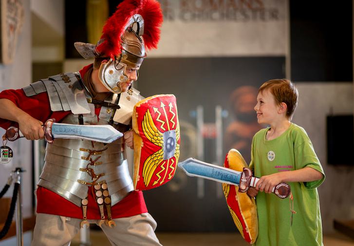 A man dressed as a Roman soldier is holding a shield and pretend sword and is challenging a young boy, also with a shield and sword, but wearing a modern green t-shirt