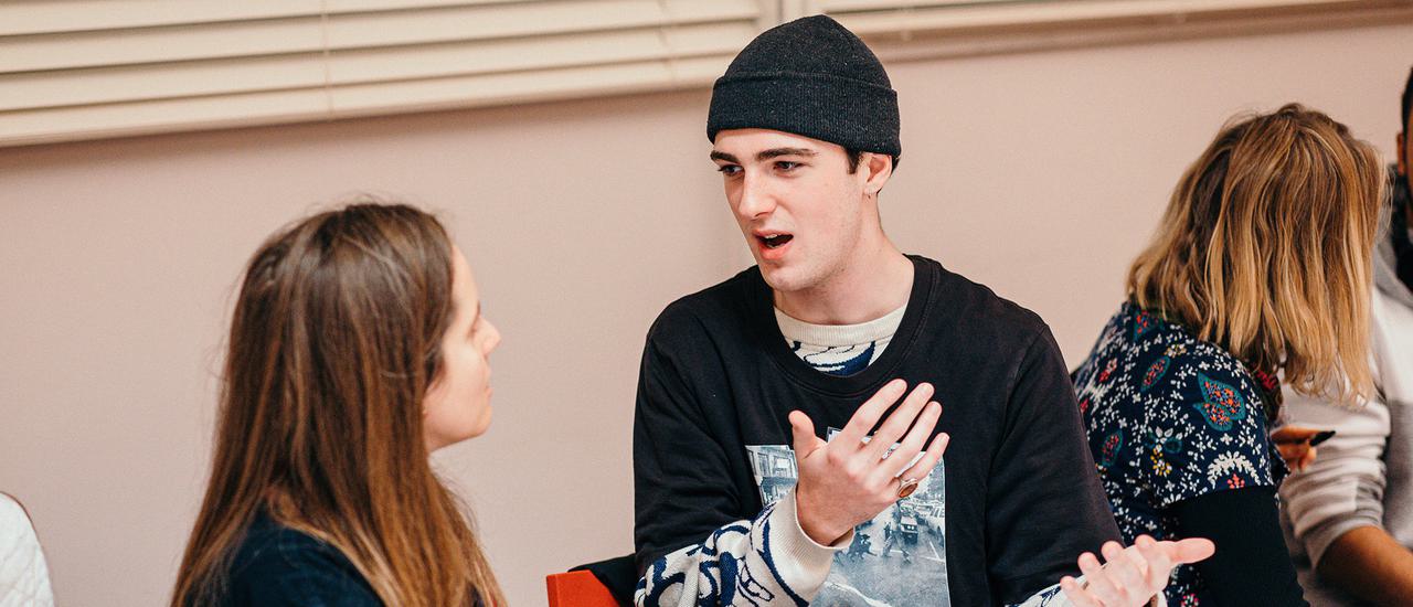 A teenage boy in a black, long-sleeved t-shirt and black beanie hat sits on a chair. He is gesturing with both his arms in front of him, and is turned to speak to a girl who sits on his right. She has long brown hair and is looking towards him.