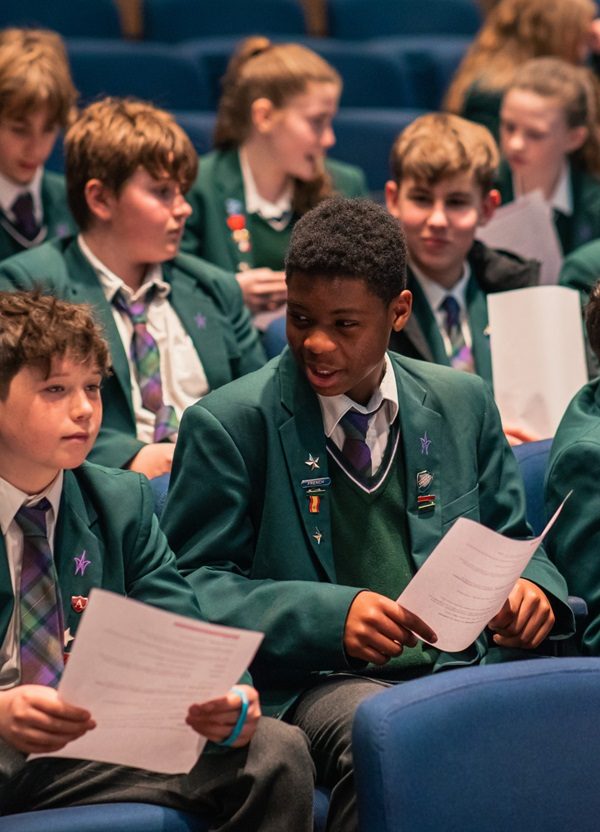 School boys dressed in green blazers are sat chatting while holding white piece of paper in a theatre auditoria.