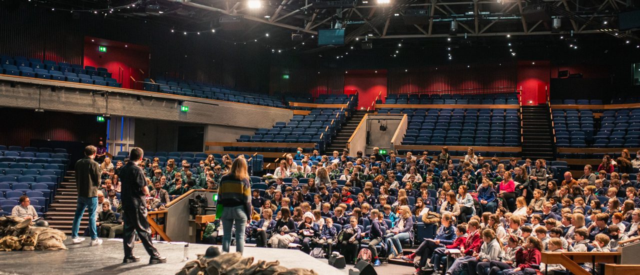 Three people on stage in front of an audience made up of different schools.