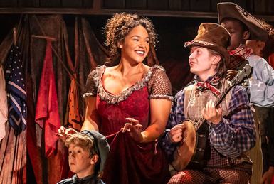 A jolly group scene of the young cast playing pretend horse and carriage. A young boy and a woman are sitting on stools on top of a table as the boy plays the banjo. The children in the scene play pretend horses in front of them in descending levels. Everyone wears colourful but well-worn clothing.