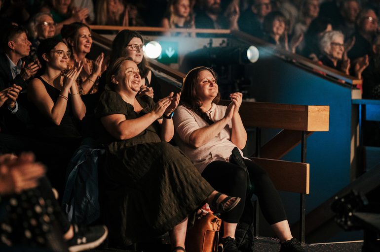 An audience in a theatre auditorium are seated and clapping with smiling faces. They're really loving the show.