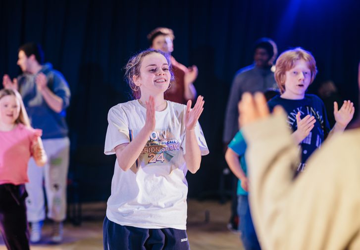 Young people perform together with a dark curtain behind them, lit in white and blue lighting. They are jumping and clapping, in the middle of a dance. The performer in the centre smiles enthusiastically and they all look like they're having a great time.