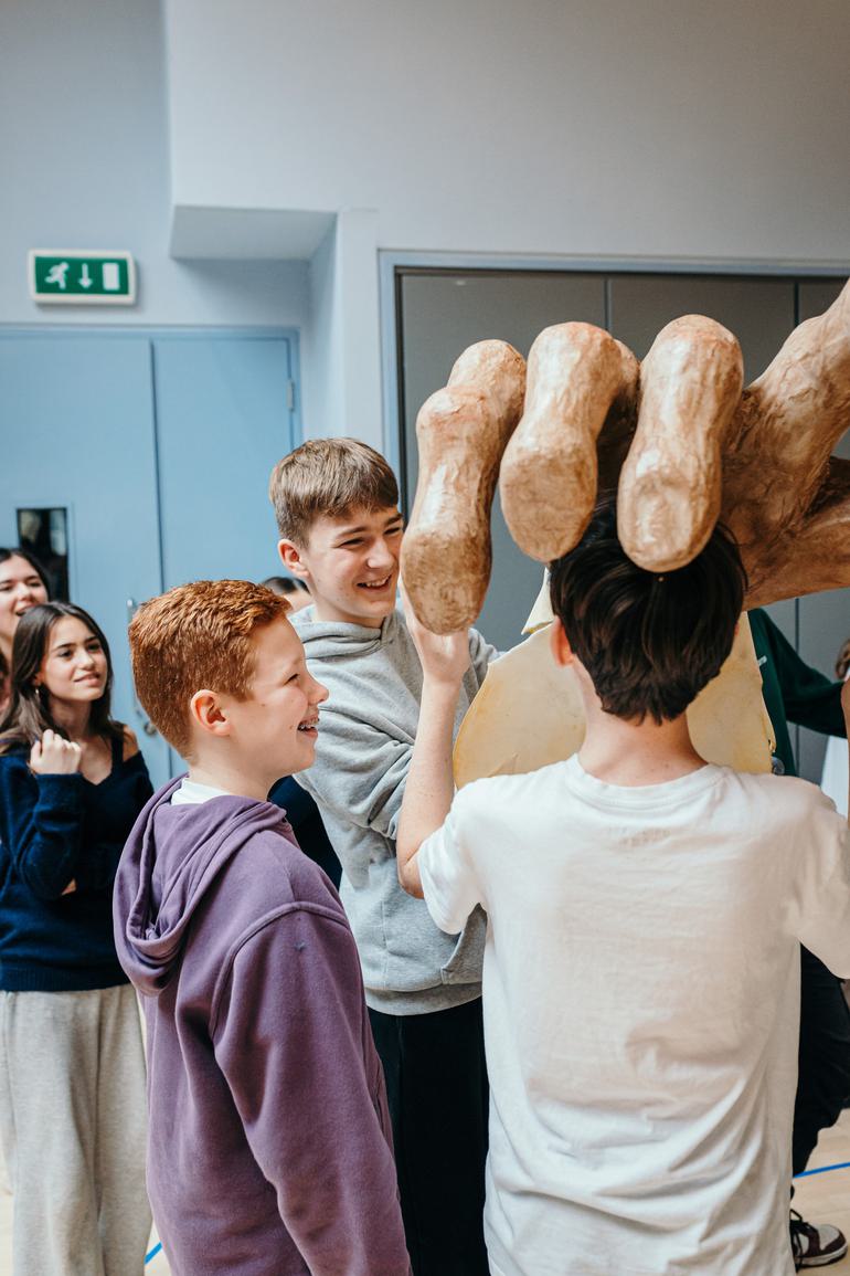 Three young performers hold up a huge oversized model hand. They are smiling and enjoying themselves. Other young performers look on from the background.