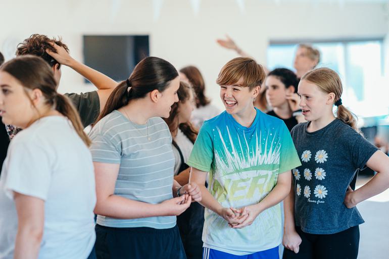 A group of young performers stand together in a rehearsal room, talking and laughing together. They look relaxed and like they are having a really great time.