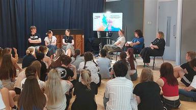 A group of Chichester College students sit on the floor infront of a panel of speakers and a TV screen.