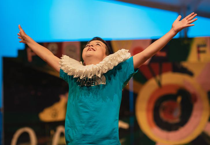 A young boy stands onstage, with a beaming smile on his face, his head thrown back and arms outstretched in a joyful pose. He is wearing an oversized Elizabethan ruff over his blue t-shirt.