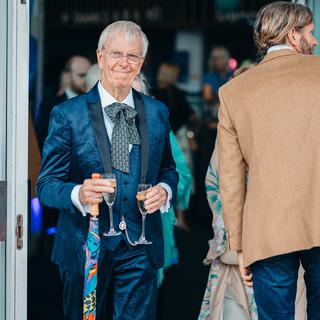 A grey-haired man in a three piece blue velvet suit and patterned blue cravat, carries an umbrella and two champagne glasses as he approaches the camera.