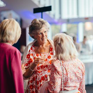 Three women chat animatedly to each other in the theatre foyer; the woman in the centre wears a party dress of orange flowers on white and carries a glass of champagne
