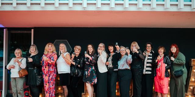 A group of people gather outside CFT Foyer smiling and waving for a picture.