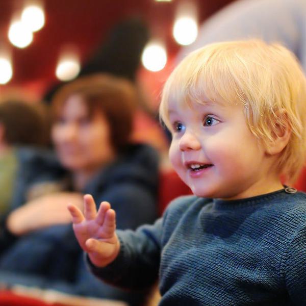 A young boy is marvelled looking at a stage. He sits in red theatre seats with sparkling lights in the background.