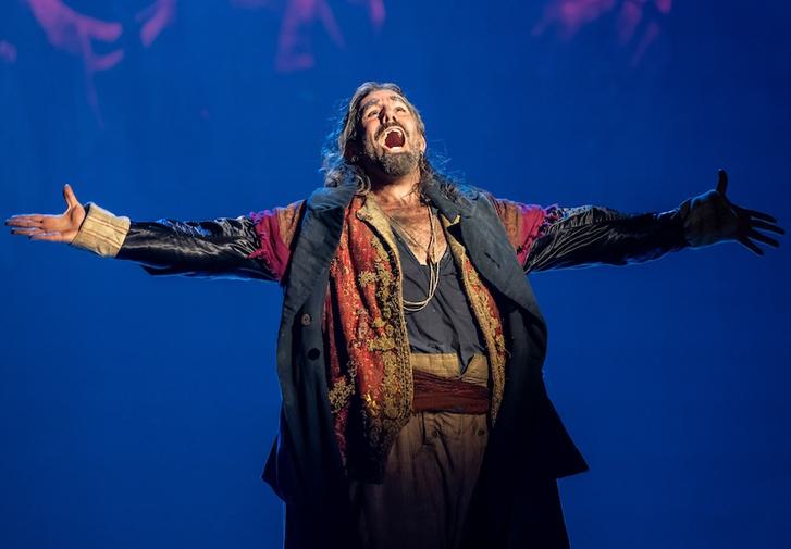 A man stands alone with his arms spread wide open in song. He has long black hair and a thick black beard. He wears a layered collection of well-worn clothes.