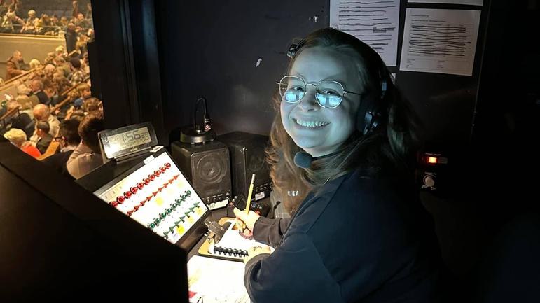 Zoe is sitting in a darkened sound booth with her face lit from below by the lights on the control panel. She has a beaming smile on her face as she looks towards the camera.