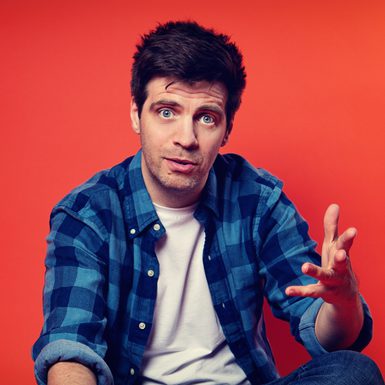 An image of Ed Patrick who is sitting looking at the camera wearing a blue check open shirt, a white t-shirt and is gesturing with his hand as if explaining something.