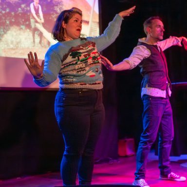An image of two performers on stage (Laura and Paul). They both look like they are doing a dance move of an arm wave in front of a projector.