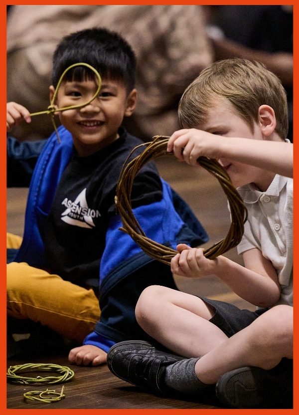 A young boy uses a makeshift willow hoop as a magnifying glass and peers through smiling.