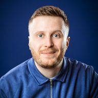 A photo of Josh Baulf looking straight to camera with a slight smile. He has short blonde/brown hair and a stubble. He is wearing a blue polo shirt.