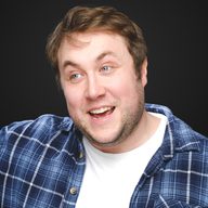 A photo of Tom Toal, looking slightly off to the side. He is an amused expression. He is wearing a white t-shirt with an open blue checked shirt over the top.