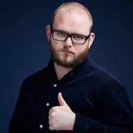 A photo of Will Duggan wearing a dark shirt posing with his thumb up. He is looking to camera, he has a bald head, dark, short beard, and is wearing glasses.