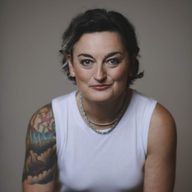 An photo of Zoe Lyons wearing a white sleeveless t-shirt and looking straight to camera with a smile. She has a tattoo on her right arm, and short dark hair.