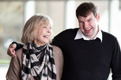 A woman and a younger man stand laughing together in the theatre foyer. She is looking up at him and he has his arm around her shoulder for support.