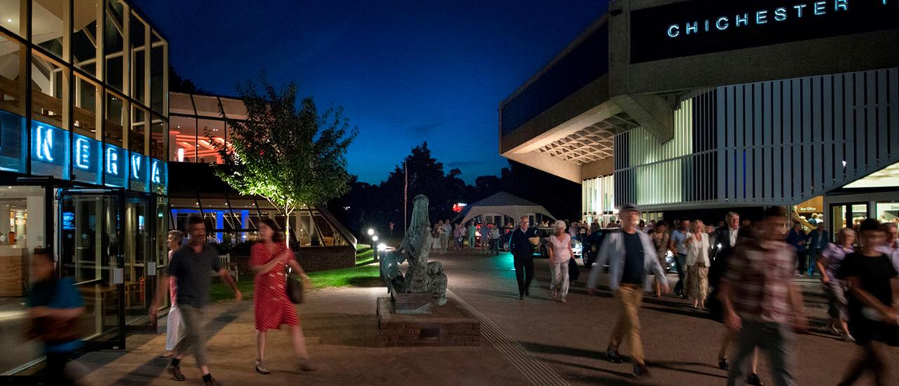 A photo of the outside of the theatres at night. Both buildings are lit up and audiences are streaming out of both the Festival and Minerva Theatre doors