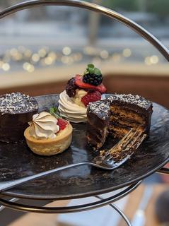 The top of three tiers of afternoon tea stand with beautiful selection of cakes; a fork has sliced the chocolate opera cake in two