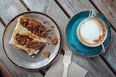 A slice of carrot cake next to a cup of frothy coffee on a wooden picnic bench.
