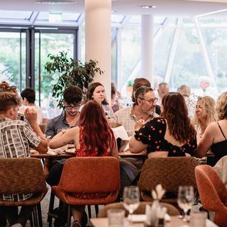 A group dining in The Brasserie on a long table. In the foreground is a blurred plant to the right, behind the long table is a backdrop of the greenery of Oaklands park out of the window.
