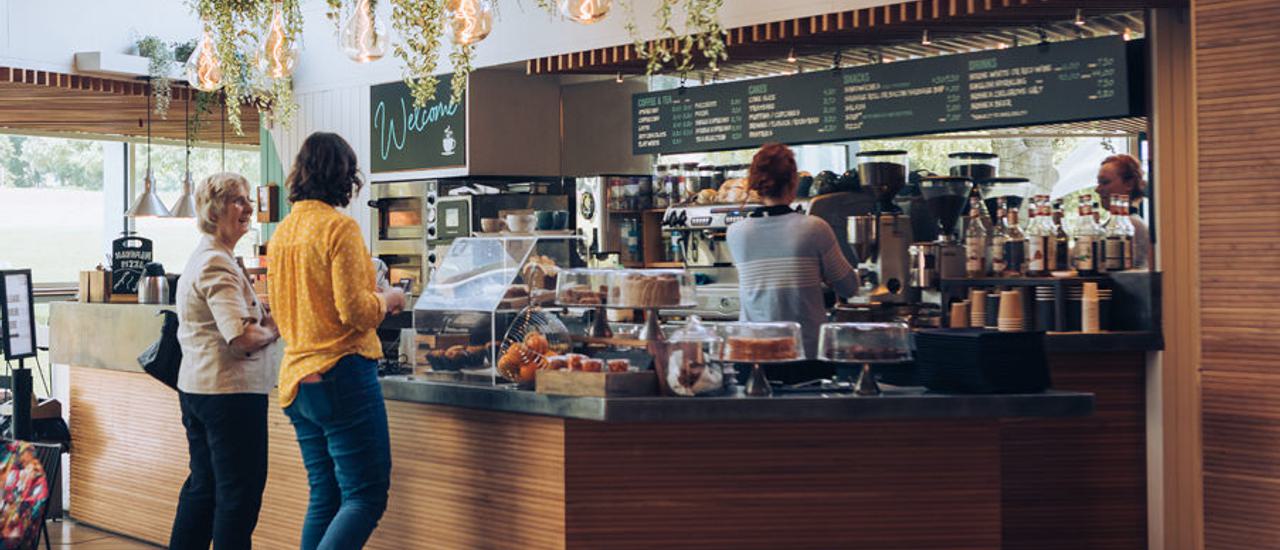 Two customers are waiting at the counter in our cafe which is loaded with delicious looking cakes. Behind the counter a staff member is making barista coffees.
