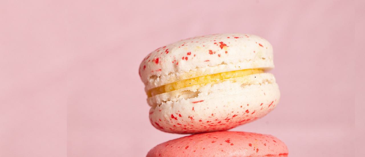 A macaron (cream with pink splodges and a cream filling) balances on a pink macaron (with darker pink splodges); the background is pale pink.