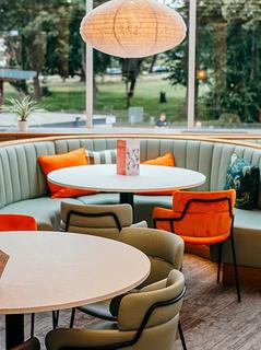 A view of the Minerva Bar, the main focus being a couple of tables one of which is a booth with colourful cushions on the seats leading to the greenery of Oaklands park out of the window.