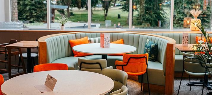A view of the Minerva Bar, the main focus being a couple of tables one of which is a booth with colourful cushions on the seats leading to the greenery of Oaklands park out of the window.