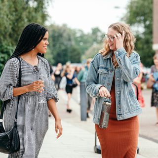 Two young women, one with long dark hair wearing a grey dress, and one with light brown hair, a denim jacket and orange dress, smile and talk as they approach the Theatre.
