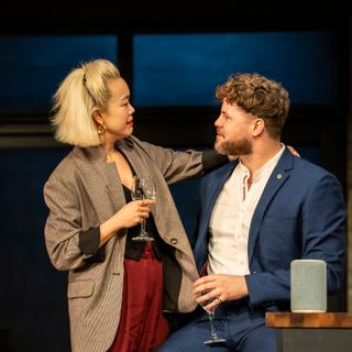 A woman with shoulder-length blonde hair stands looking at a bearded, brown-haired man who's sitting next to her. She has her arm around his shoulders and is holding a wine glass in her other hand, and is wearing a brown jacket, red trousers and gold hoop earrings. The man is turned to face her and is also holding a wine glass, wearing a white shirt and blue suit.