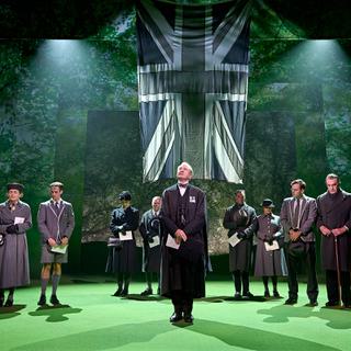 The company of 11 actors, all wearing dark, sombre clothing, stand with their heads bowed. In the centre, Julian Wadham, wearing a number of wartime medals, stands looking up; above them is a grey Union Jack  with the colour drained out. The floor is a vivid green and behind them are receding backdrops of verdant trees and leaves.