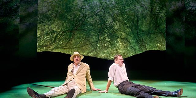 Rupert Everett, in white shirt and trousers and beige jacket, sits on the ground with his legs stretched out in front of him, next to Jack Bardoe who's in white shirt and dark trousers. The ground is green and behind them is a backdrop with light filtering through green trees and leaves.