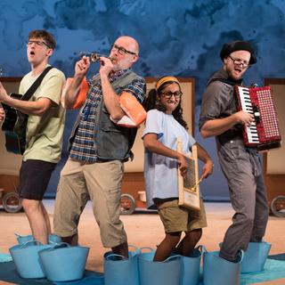 Three people stand on stage, with their feet in big, blue plastic buckets. They are all playing instruments. On the left, a young man in a green t-shirt and white shorts strums a black guitar. In the centre, an older man plays a tiny trombone and a young woman plays a washboard. On the right, a man in a black hat plays a red accordian. They have focused faces, and are playing enthusiastically.