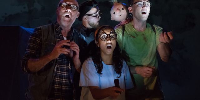 Four actors stand on a dark stage, holding torches up to their faces. It looks mysterious. In the centre is a young woman in a white t-shirt, to the left is an older man in a blue checked shirt. On the right is a young man in a green t-shirt, with a baby puppet on his back. At the back is a man in a beret, who is looking at the baby. They are all wearing glasses.
